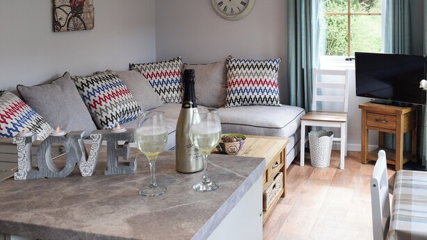 Een gezellige woonkamer met een bank, een bijzettafeltje met glazen en een fles, een televisie op een houten dressoir, en een klok aan de muur.