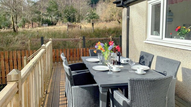 An outdoor dining area with a table set for two, surrounded by wicker chairs, overlooking a mountainous landscape.