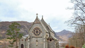A stone church with a prominent rose window and a bell tower, surrounded by a grassy area and mountains in the background.