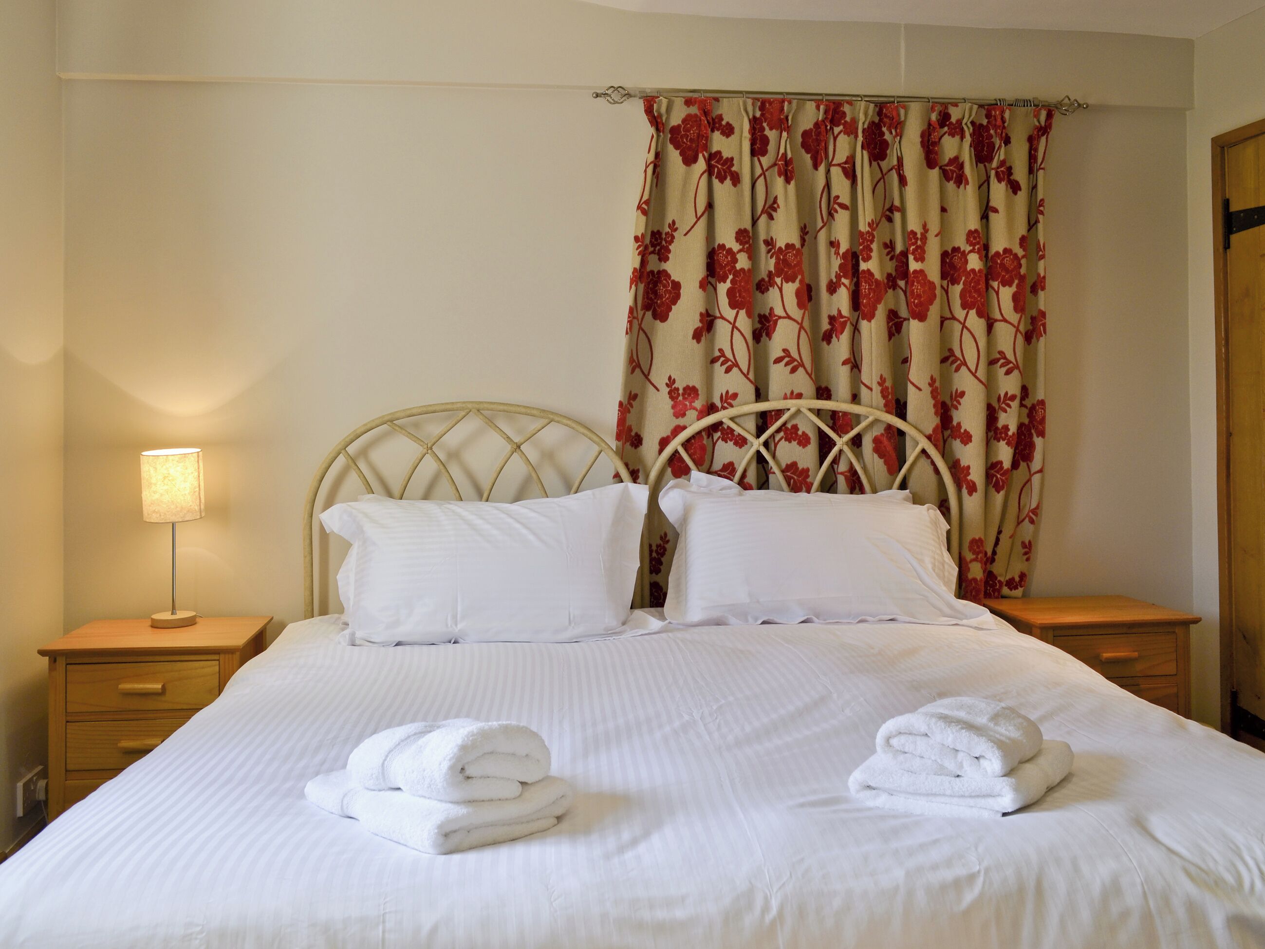 A neatly made bed with white linens and two rolled towels, a bedside lamp, and a floral curtain.