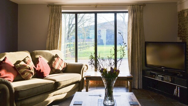 A living room with a sofa, a coffee table with a vase, a flat-screen TV, and a large window with curtains. - 4 bedroom accommodation in Uggeshall, near Southwold (Uggeshall, near Southwold)