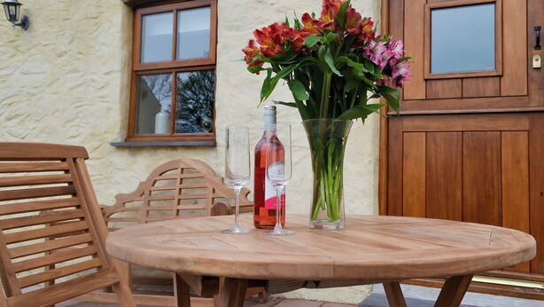 A round wooden table with a clear glass bottle and a glass of red wine, a vase with flowers, and wooden chairs in front of a stone building with windows and a wooden door. - 3 bedroom accommodation in Llanynghenedl, near Holyhead (Llanynghenedl, near Holyhead)