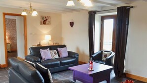 A living room with a black leather sofa, a purple coffee table, and a black armchair. There is a window with dark curtains, a wall-mounted lamp, and a small table with a lamp and figurines. - 3 bedroom accommodation in Llanynghenedl, near Holyhead (Llanynghenedl, near Holyhead)