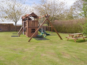A wooden play structure with a slide and swings, a picnic table, and a brick wall in the background.