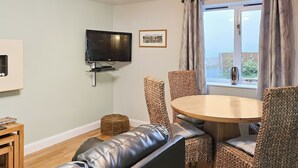 Een woonkamer met een leren bank, een houten eettafel met gestreept zittende stoelen, een aan de muur bevestigde televisie en een raam met gordijnen.