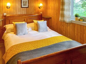 A wooden-paneled room with a bed, two wall-mounted lamps, a window with a view, and a small shelf with plants.