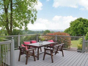 A deck with a table and chairs, surrounded by a railing, overlooking a landscape with trees and hills.