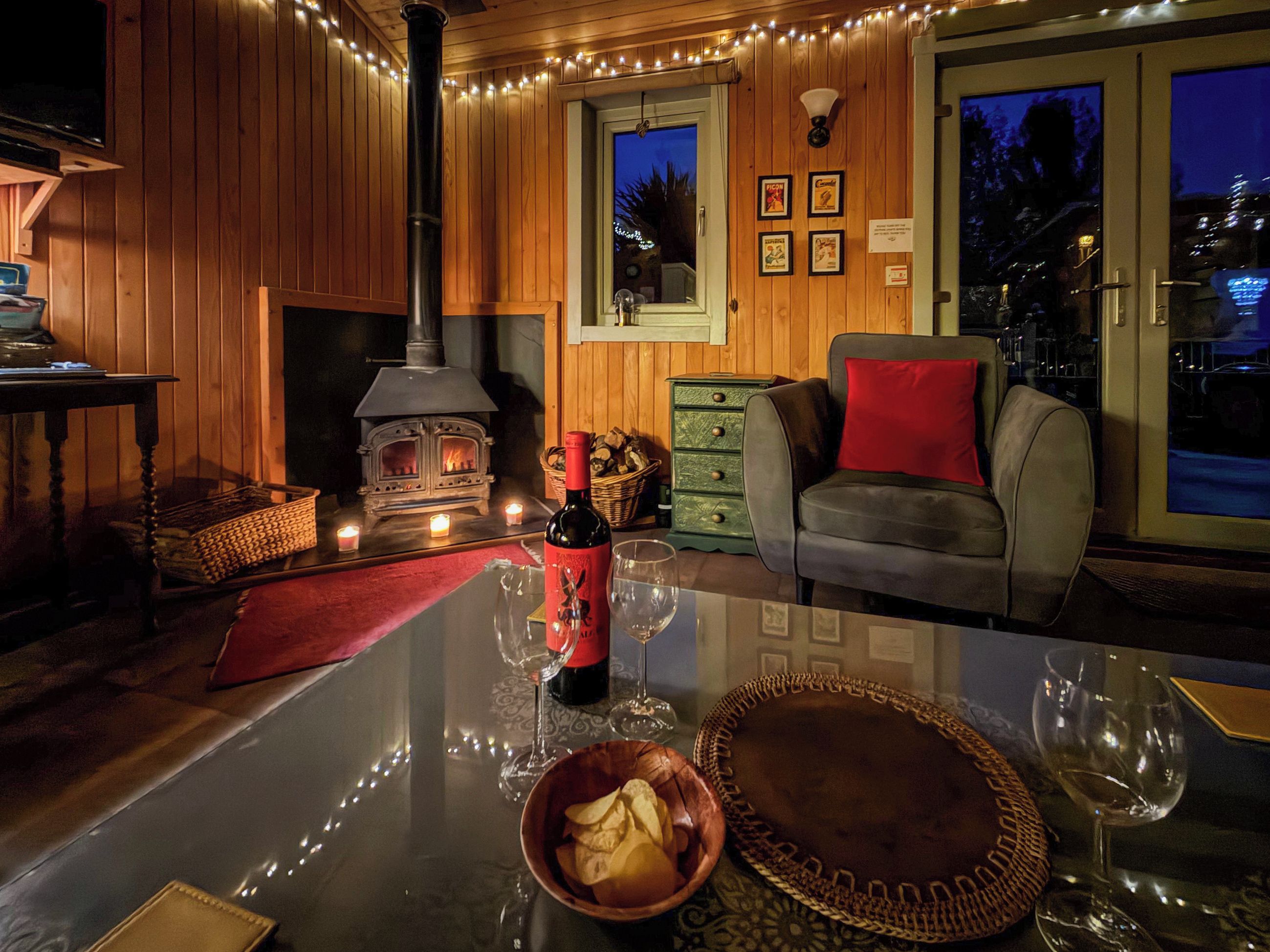 A cozy room with a wood-burning stove, a sofa, a dining table with wine glasses, and a view of the outside.
