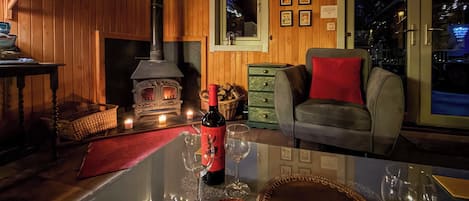A cozy room with a wood-burning stove, a sofa, a dining table with wine glasses, and a view of the outside.