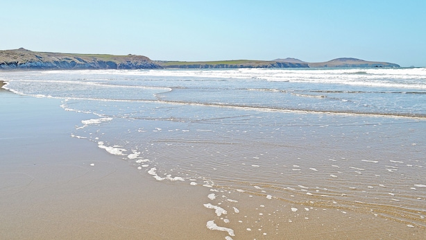 Ein Strand mit Wellen, Sand und sanften Hügeln im Hintergrund.