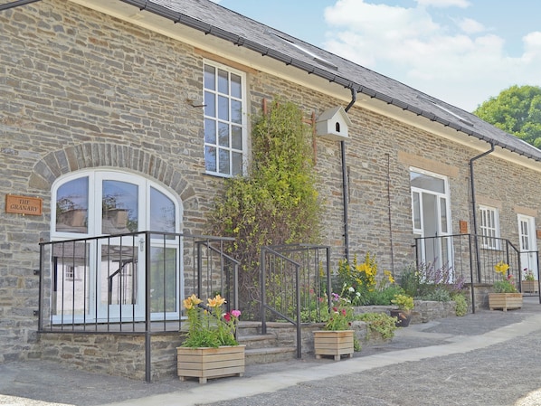 A stone building with a sign reading "The Granary." It features a balcony with a black railing, wooden flower planters, and a small birdhouse on the roof.
