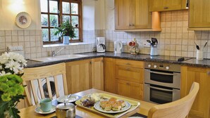 A kitchen with wooden cabinets, a sink, a window with curtains, and a dining table set with plates, cups, and a teapot. - Cider Cottage - E5576 (Chittlehampton, near Umberleigh)