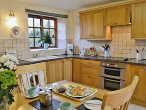 A kitchen with wooden cabinets, a sink, a window with curtains, and a dining table set with plates, cups, and a teapot. - Cider Cottage - E5576 (Chittlehampton, near Umberleigh)