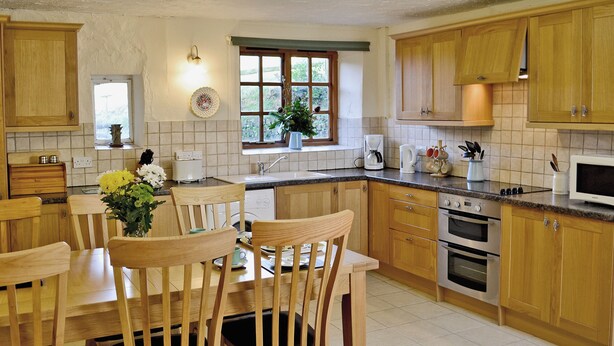 A kitchen with wooden cabinets, a dining table with chairs, a window with a view, and a microwave. - Cider Cottage - E5576 (Chittlehampton, near Umberleigh)