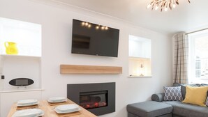 A modern living room with a fireplace, a flat-screen TV, and a dining area with plates and cutlery. - One Beckside Cottage (Staithes, near Whitby)