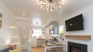 A modern living room with a sofa, a dining table with benches, a fireplace, and a kitchen in the background. - One Beckside Cottage (Staithes, near Whitby)
