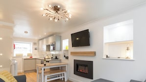 A modern living room with a fireplace, a television mounted on the wall, and a kitchen area with white cabinets and a wooden bench. - One Beckside Cottage (Staithes, near Whitby)