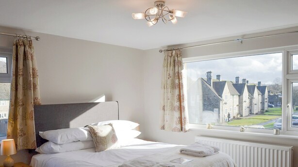 Un dormitorio con un ventanal grande, una cama con ropa blanca, una mesita de noche con lámpara, y vistas a casas afuera.