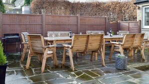 Un patio al aire libre con mesa y sillas de madera, una parrilla y piso de baldosas de piedra.