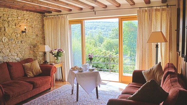 A cozy room with a stone wall, wooden beams, a red sofa, a table with a vase of flowers, and a balcony with a view of greenery. - 4 bedroom accommodation in Colyton (Colyton)