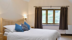 A neatly made bed with white bedding and blue pillows, a wooden headboard, a nightstand, and a window with curtains. - 4 bedroom accommodation in Colyton (Colyton)