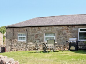 Ein Steinhaus mit einem Ziegeldach, zwei Fenstern, einem Grill und einem Tisch mit Stühlen im Hof.
