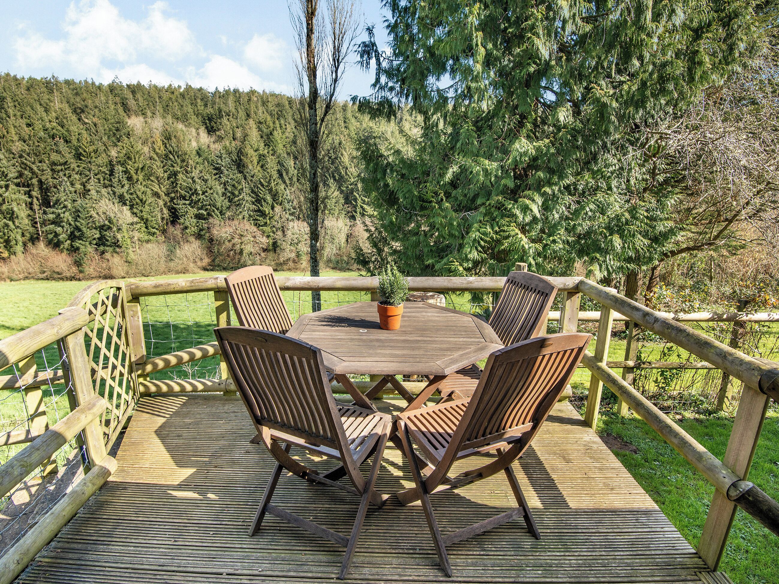 Une terrasse en bois avec une table ronde et quatre chaises, entourée d’une balustrade en bois, donnant sur une zone boisée.
