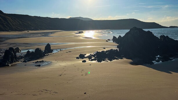 Uma praia com céu claro, sol brilhando e formações rochosas perto da costa.