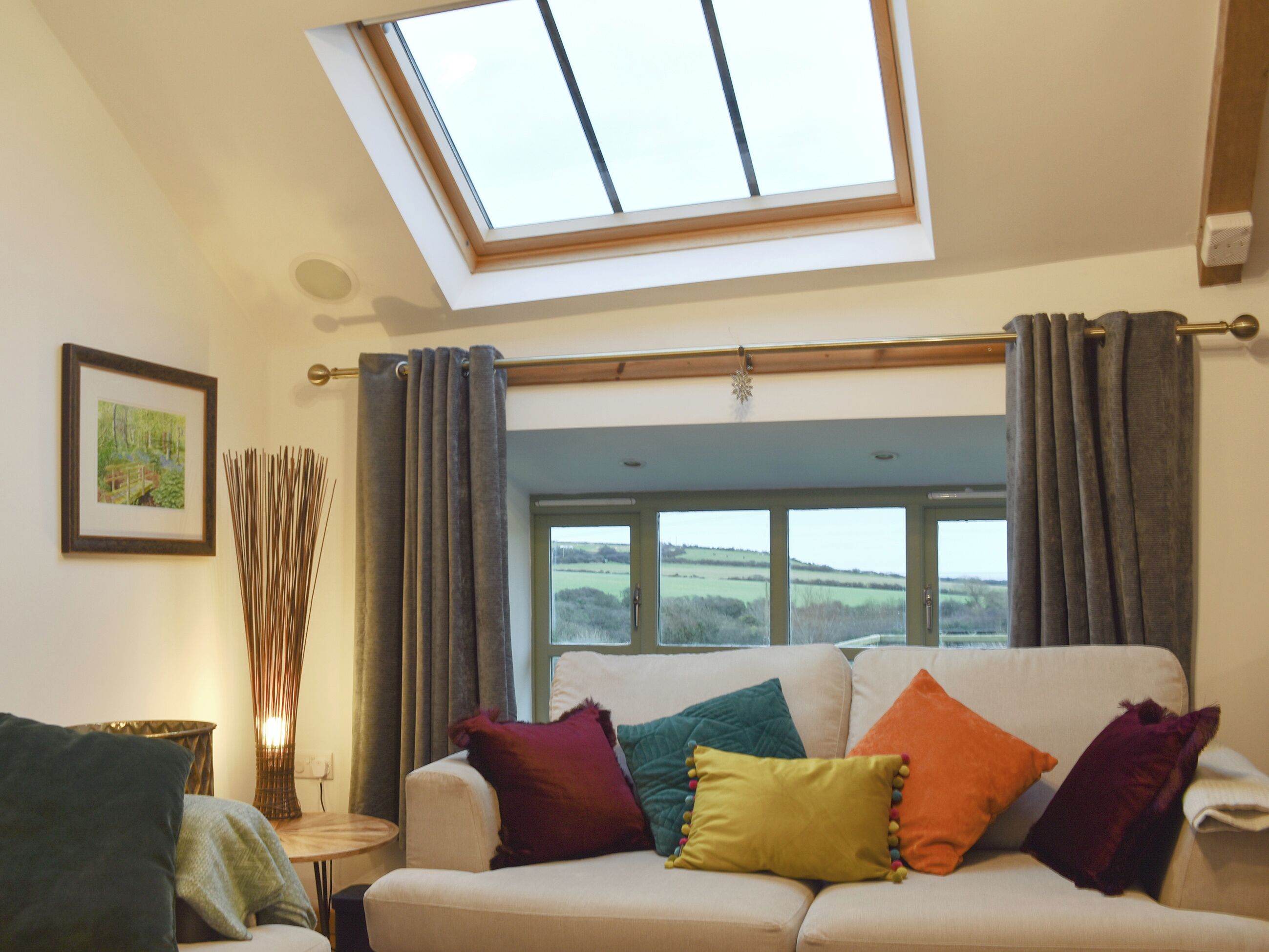 A living room with a skylight, a sofa with colorful cushions, a framed picture on the wall, and a window with a view of the countryside.