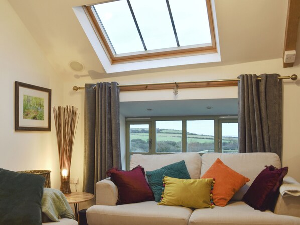 A living room with a skylight, a sofa with colorful cushions, a framed picture on the wall, and a window with a view of the countryside.
