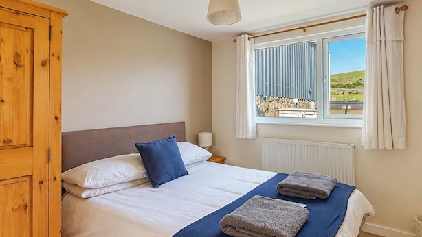 A bedroom with a bed, pillows, a wooden wardrobe, a window with curtains, and a view of the outside. - 2 bedroom accommodation in Llanllechid, near Bangor (Llanllechid, near Bangor)