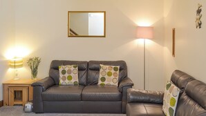 Ein Wohnzimmer mit braunem Ledersofa, zwei passende Kissen, einem kleinen Holztischchen mit einer Lampe, einer Stehlampe und einem Spiegel an der Wand.