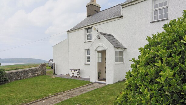 A white house with a stone wall, a small table, and a green bush in the foreground. - 1 bedroom accommodation in Cilan, near Abersoch (Cilan, near Abersoch)