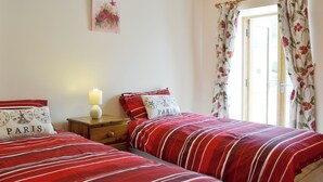 A bedroom with two beds, a nightstand, a lamp, a door with curtains, and a wall with a floral painting. - 2 bedroom accommodation in Carmarthen (Carmarthen)