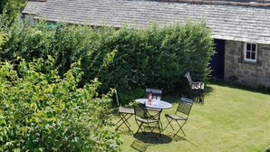 A stone building with a tiled roof, a small table with chairs, and a hedge in the foreground. - 2 bedroom accommodation in Charlestown, St Austell (Charlestown, St Austell)