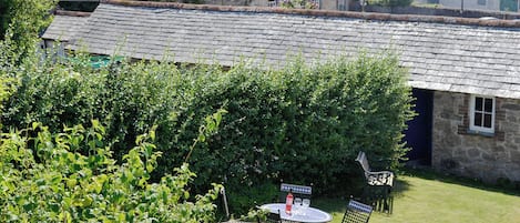 A stone building with a tiled roof, a small table with chairs, and a hedge in the foreground.