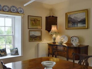 A dining area with a wooden table, chairs, a radiator, a lamp, and wall-mounted decorative plates and paintings. - 1 bedroom accommodation in Sorn, near Ayr (Sorn, near Ayr)