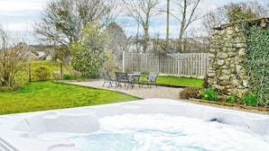 Un jacuzzi en un patio trasero con terraza de madera, pared de piedra y jardín con sillas y plantas.
