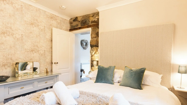 Un dormitorio con una cama grande, ropa de cama blanca y esponjosa, una pared de acento de piedra, un espejo y una mesita de noche con lámpara.