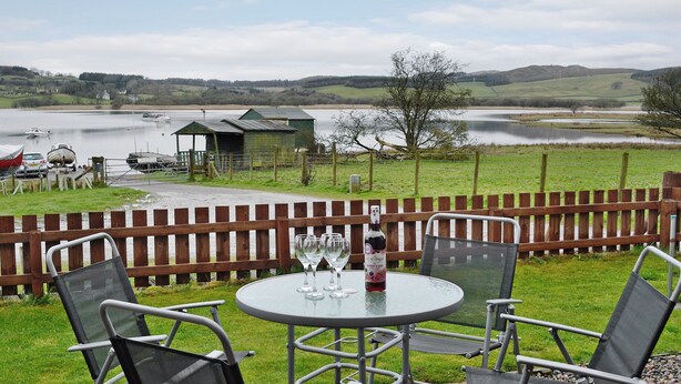 A table with chairs and a bottle of wine, overlooking a lake with boats and a wooden structure in the background.