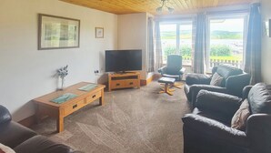 A living room with a wooden ceiling, a television on a wooden stand, a sofa, a chair, and a view through large windows.