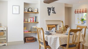 Un coin repas avec une table dressée pour quatre personnes, entourée de chaises en bois. Une bibliothèque contenant divers objets et une carte encadrée sont visibles sur la gauche.