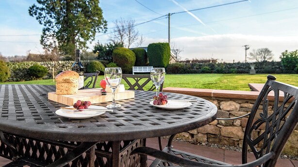A round metal table with a loaf of bread, cheese, grapes, and wine glasses set for a meal.