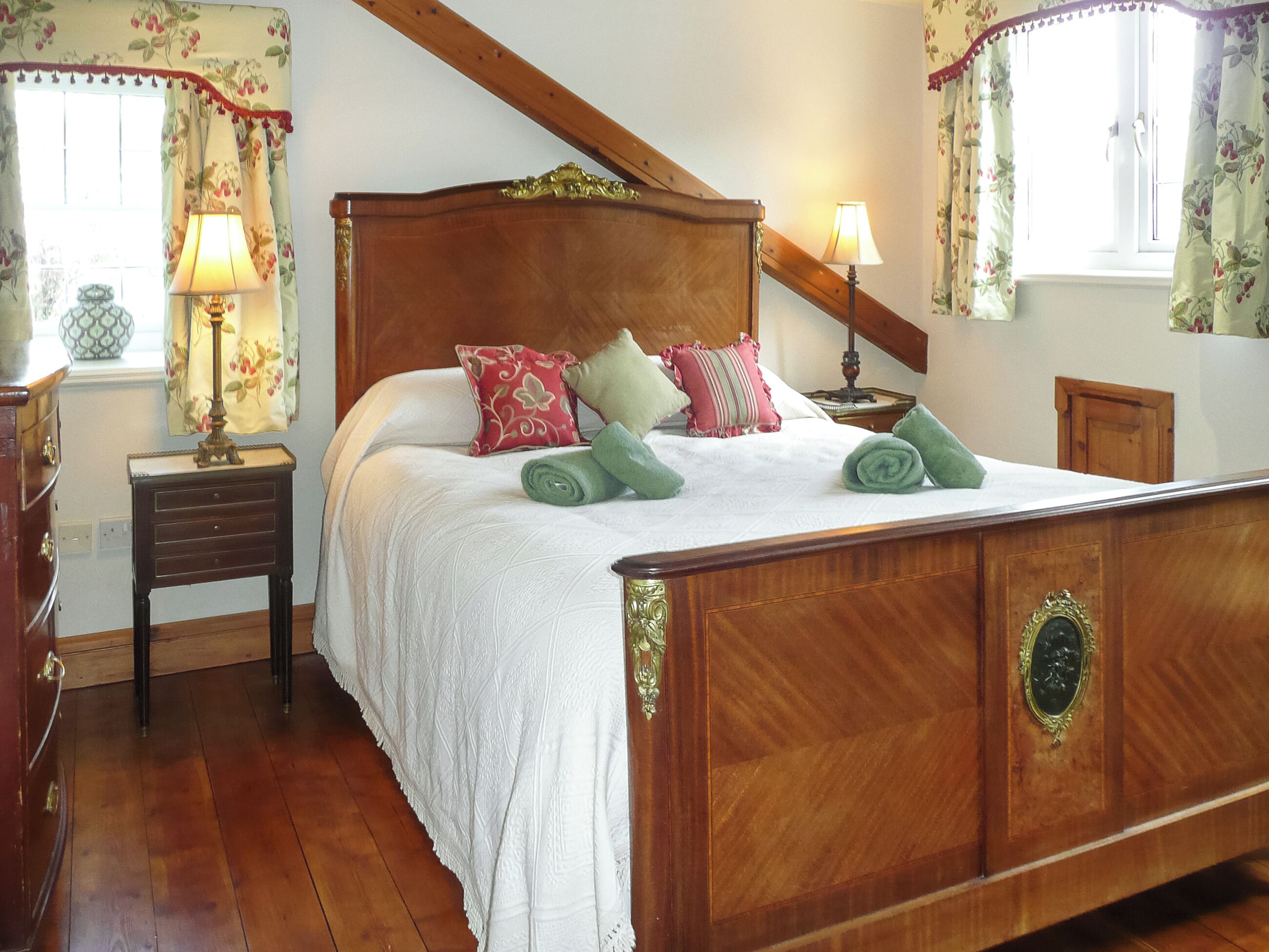 A wooden four-poster bed with white linens, multiple pillows, and green towels. There are two bedside tables with lamps, a wooden dresser, and a window with floral curtains.