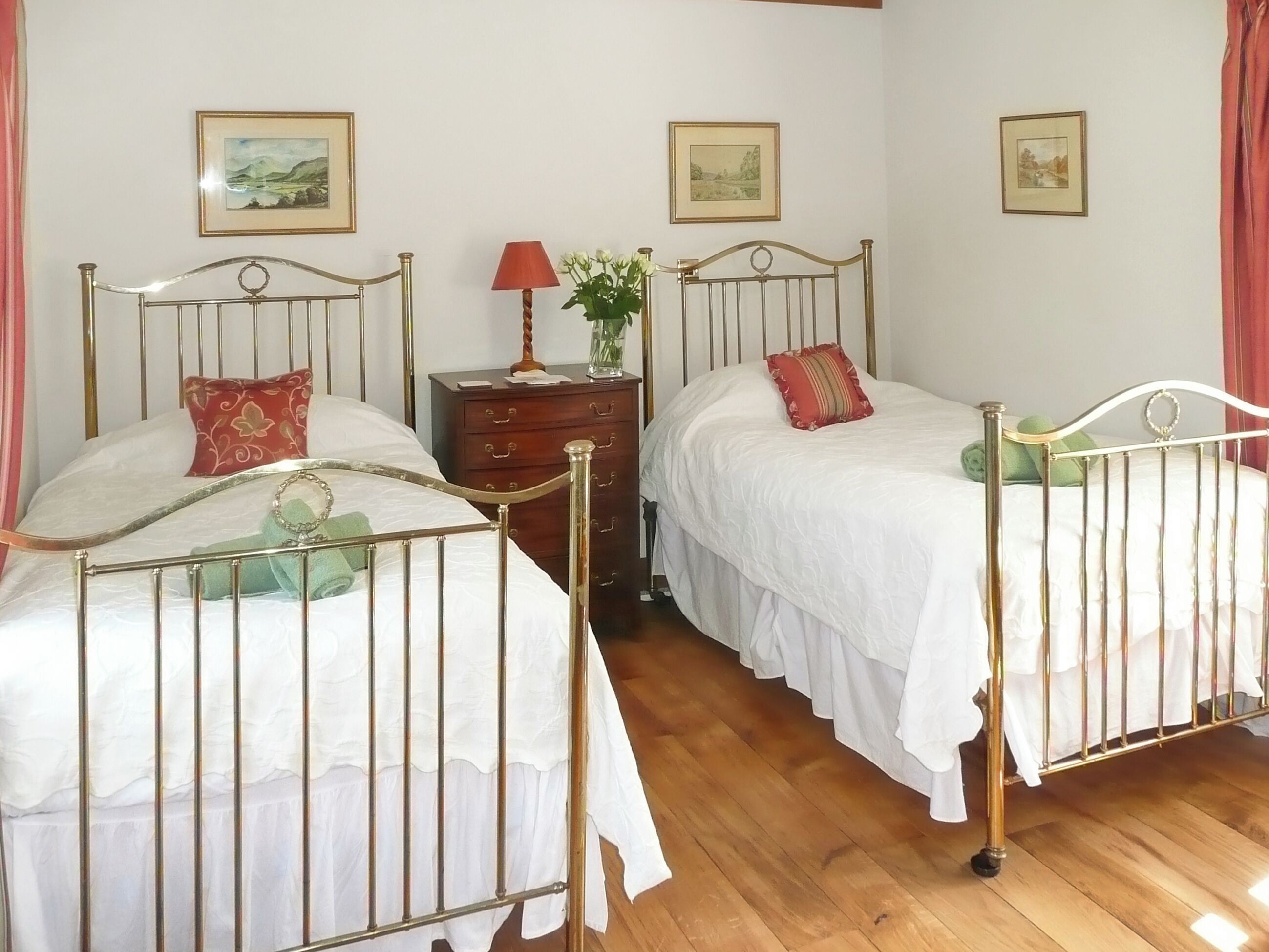 A room with two single beds, wooden flooring, a nightstand with a lamp, and framed pictures on the wall.