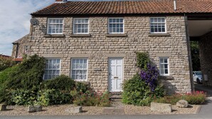 A stone building with a tiled roof, multiple windows, a white door, and a small garden with various plants and flowers.