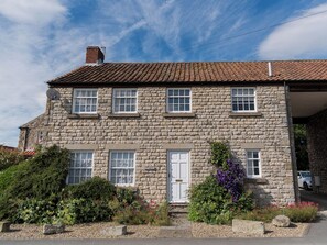 A stone building with a tiled roof, multiple windows, a white door, and a small garden with various plants and flowers. - 3 bedroom accommodation in Pickering (Pickering)