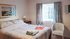 Un lit bien fait, recouvert d’une couvre-lit à motifs floraux, une chaise, une table de chevet avec une lampe et une fenêtre avec des rideaux.