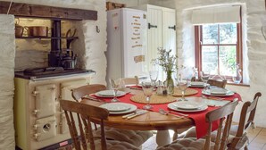 Ein Essbereich mit einem Holztisch, gedeckt für vier Personen, umgeben von Stühlen. Im Hintergrund sind ein weißer Kühlschrank und ein Herd mit Töpfen zu sehen.