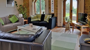 A living room with a sofa, a coffee table, and a television. There are windows with shutters and a view of the outdoors.